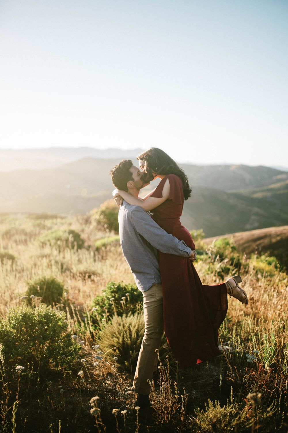 A couple sharing an intimate moment while standing in tall grass on a mountain overlook.