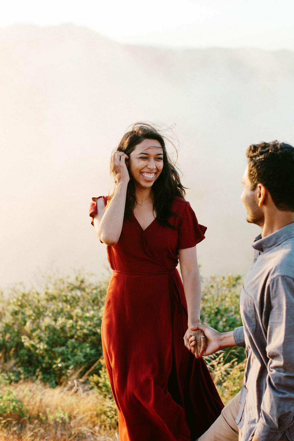 A woman in a flowing red dress smiles while standing on a mountain overlook.