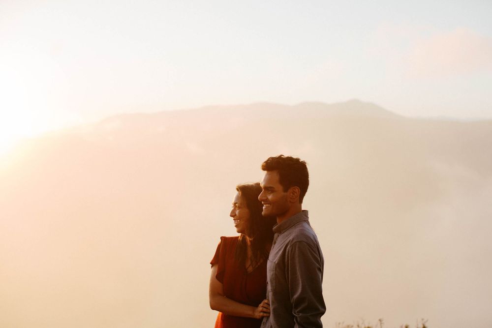A couple stands silhouetted against mountain peaks during sunset.