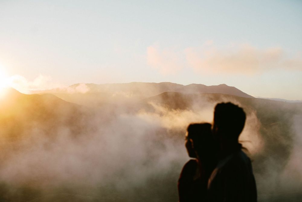 Two silhouettes stand together watching sunset over foggy mountains.