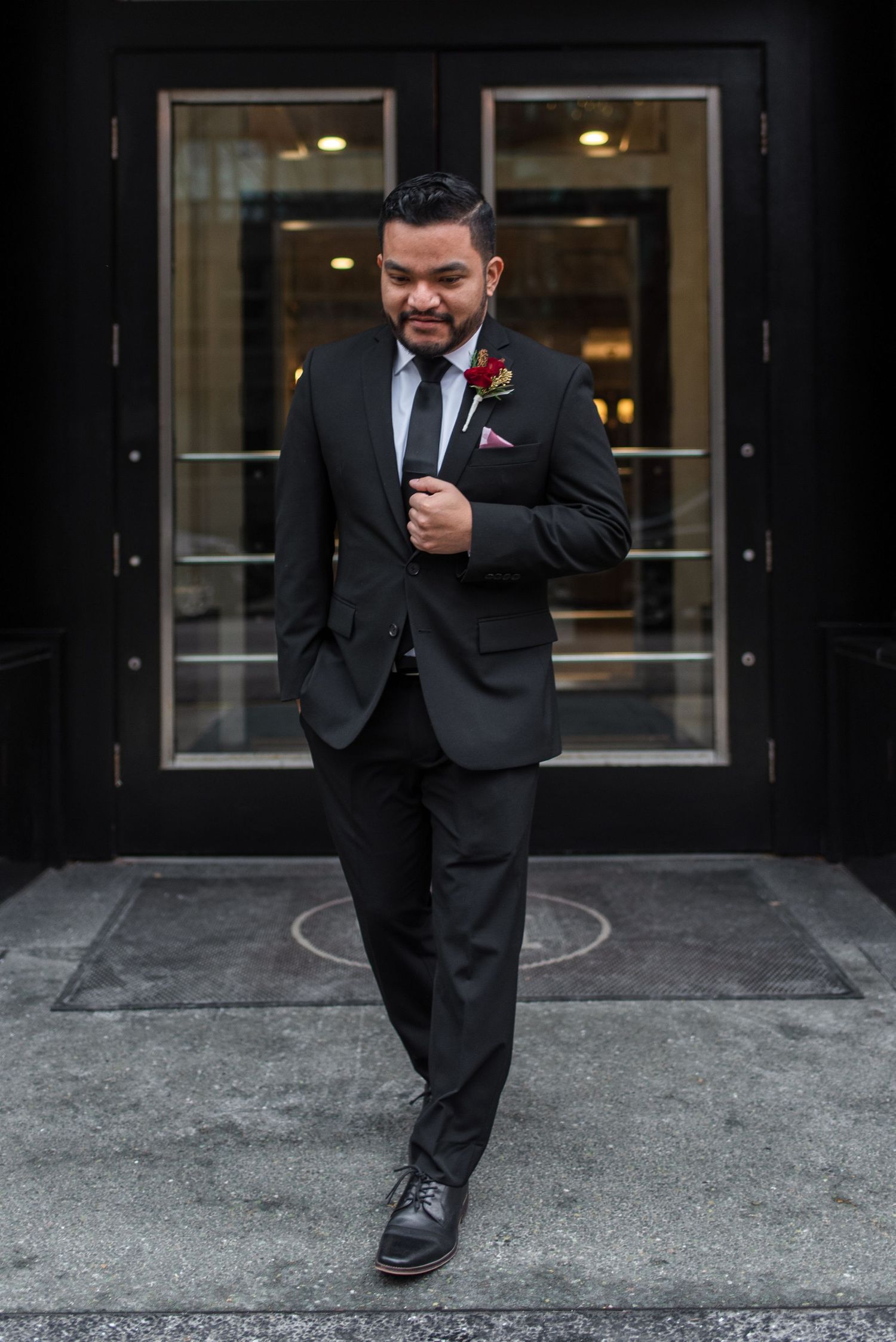 A dapper groom in a black suit and tie poses confidently outside glass doors.