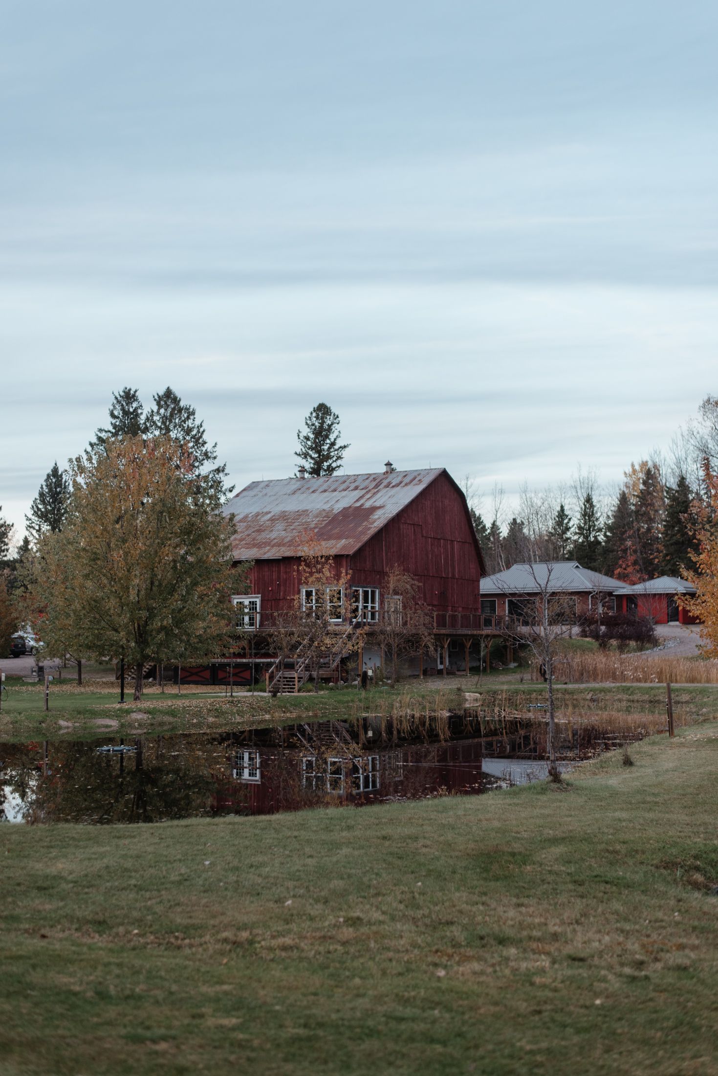Red barn reflecting in still pond water surrounded by fall foliage and evergreen trees.