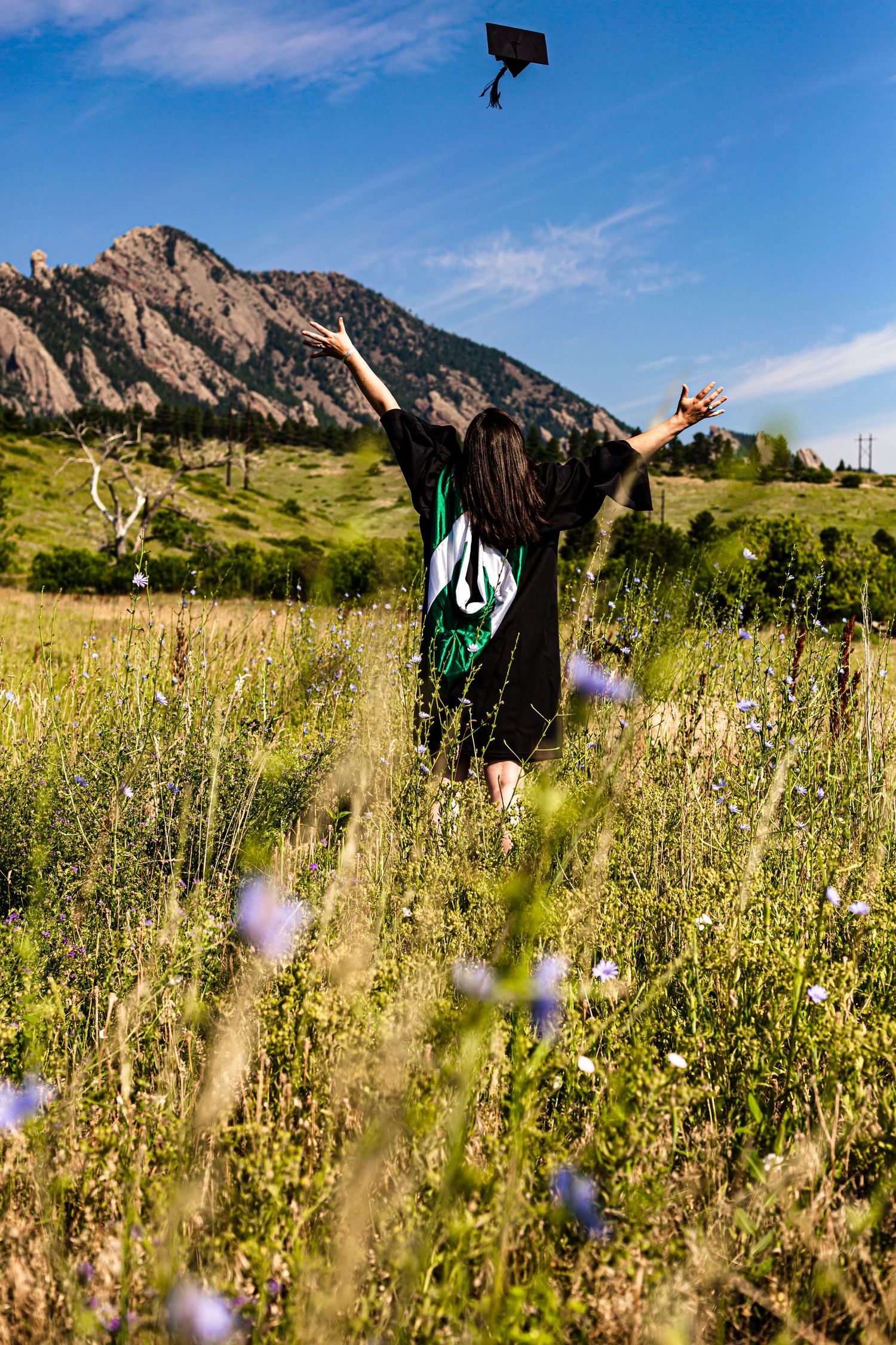 Graduate throwing cap in meadow with Flatirons mountains of Boulder Colorado in background.