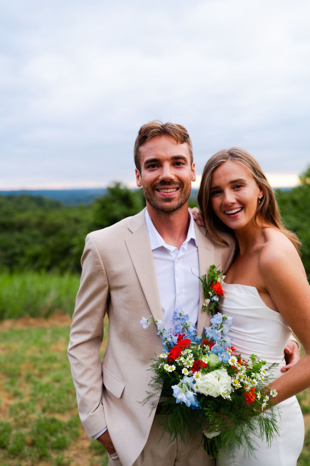 Newlyweds pose together outdoors with colorful bouquet against green landscape.