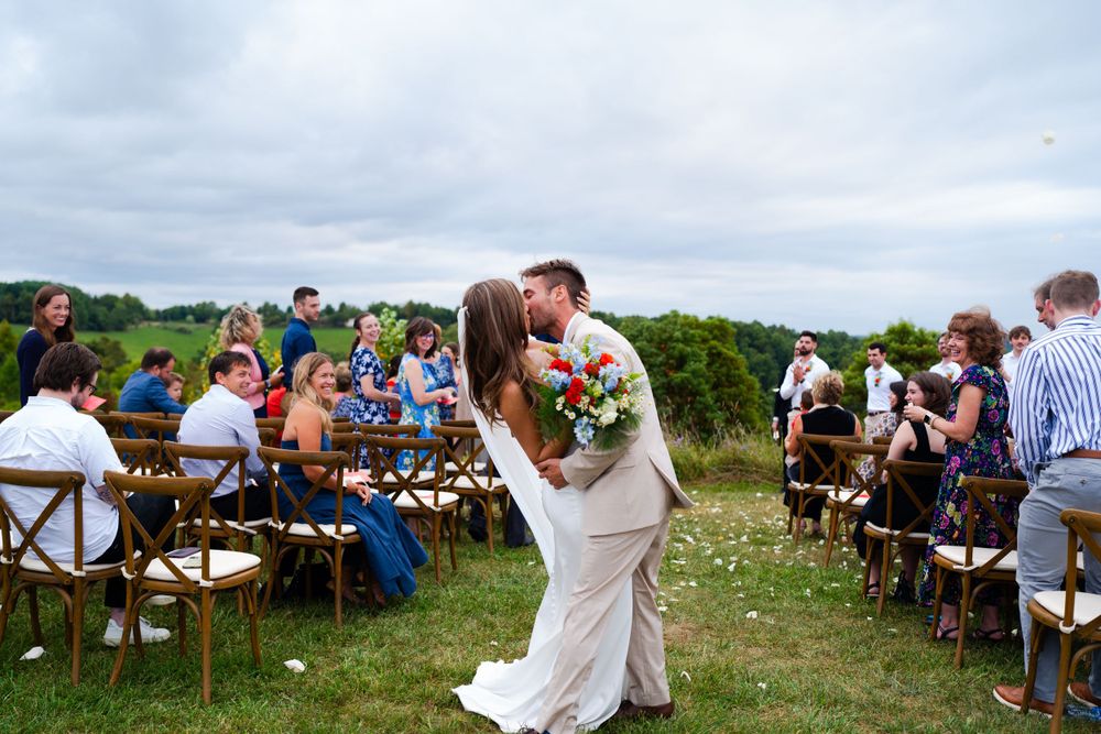 An outdoor wedding ceremony with guests seated on wooden chairs in a hilltop meadow setting.