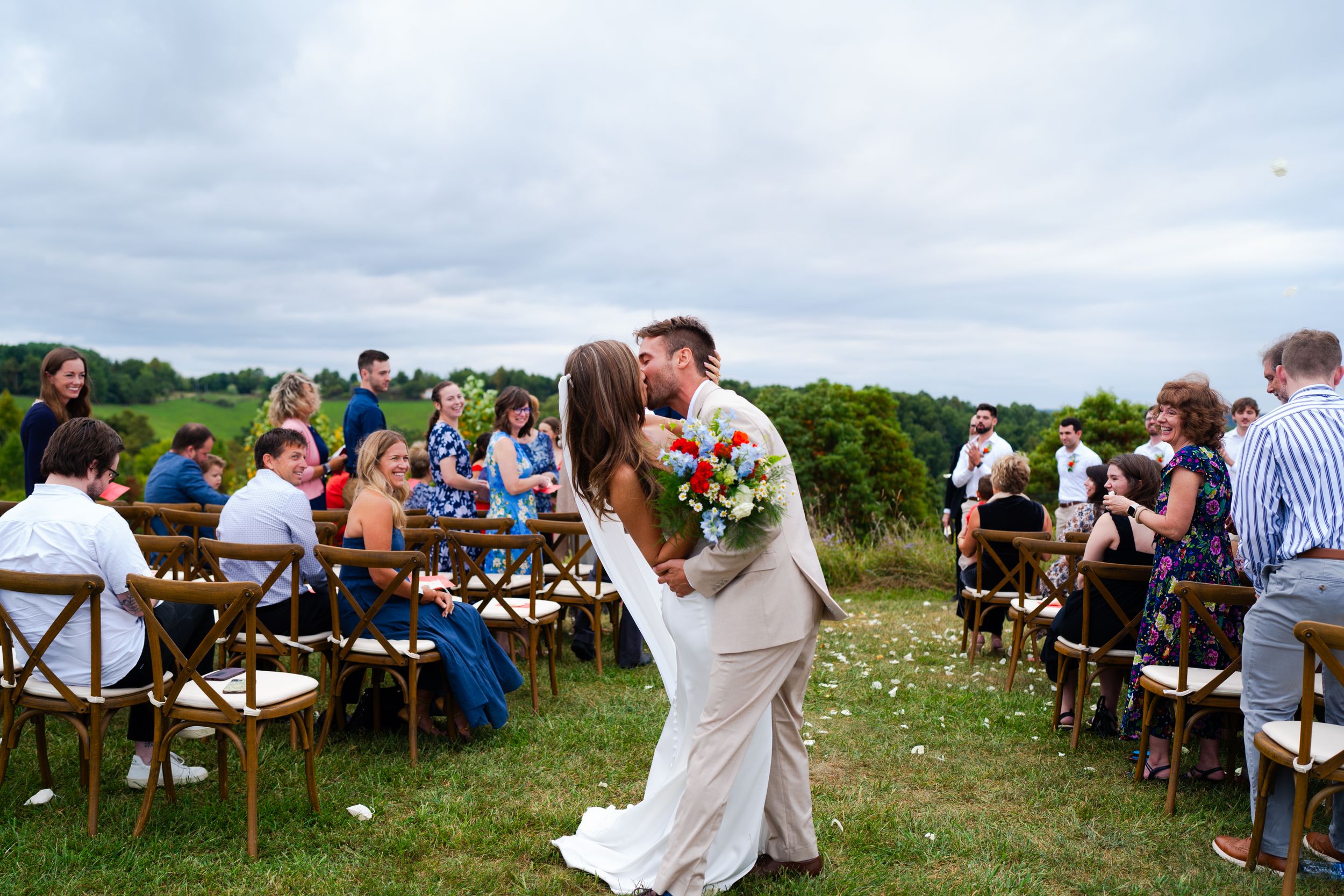 An outdoor wedding ceremony with guests seated on wooden chairs in a hilltop meadow setting.