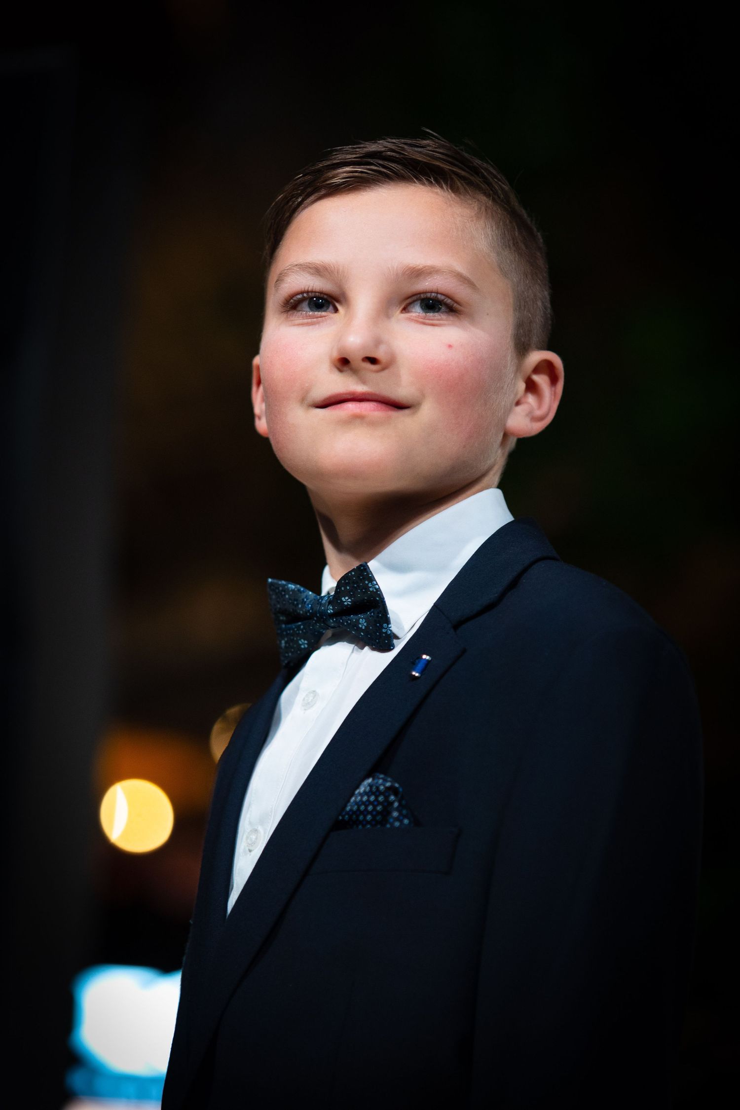 Young boy in navy blue tuxedo with bow tie and pocket square smiling at a formal event.