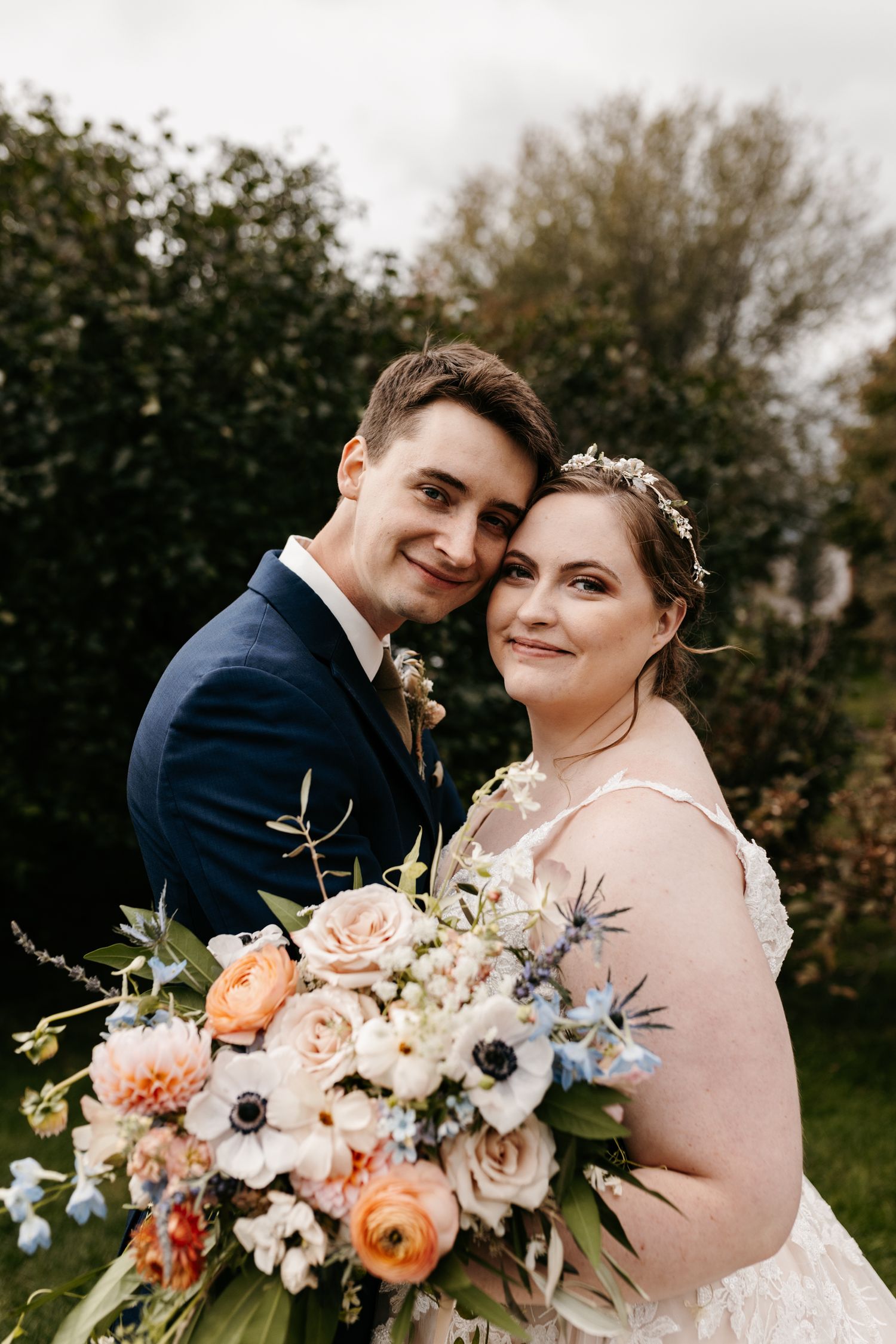 A bridal couple poses outdoors with a stunning bouquet featuring peach and white flowers.