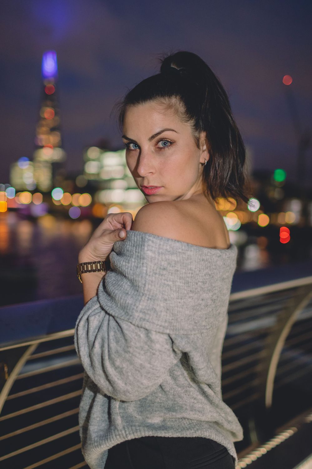White young adult female in a grey sweater posing on  millennium bridge in London with a London skyline at night