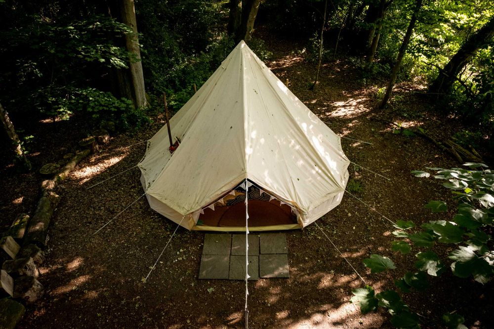 Aerial view of a beige tent in a forest clearing, surrounded by trees and stone path, under dappled sunlight.