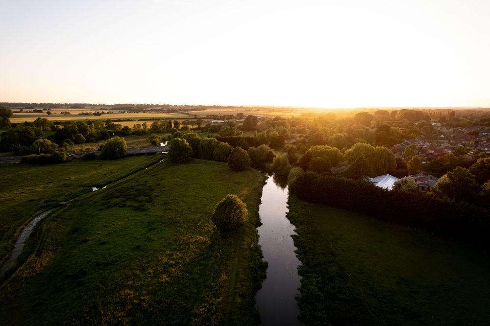 Aerial view of a peaceful countryside with a river winding through green fields and trees at sunset, with a golden sky on the horizon.