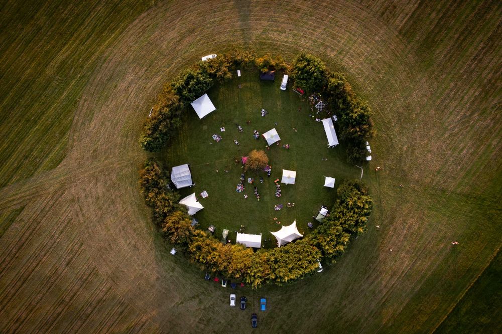 Aerial view of a circular gathering with tents and people surrounded by trees in a grassy field.