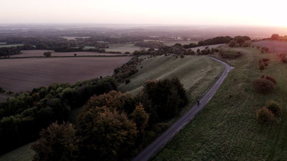 Aerial view of a cyclist on a rural road at dawn, with fields and trees on rolling hills under a soft, sunrise-lit sky.