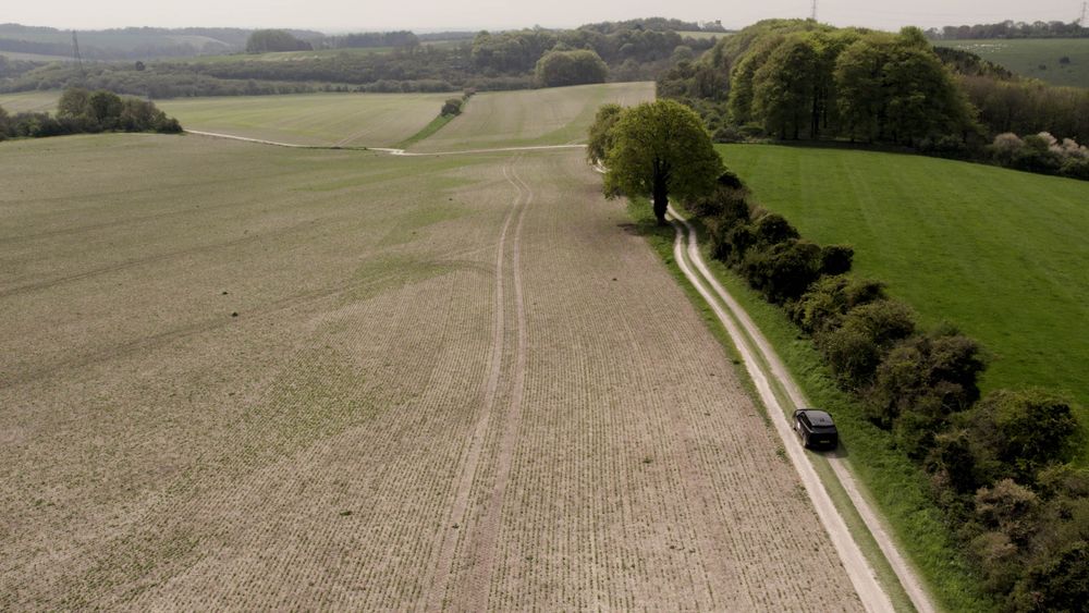 Aerial view of a rural landscape, a winding dirt road, a single tree, and a black Range Rover driving along the road under cloudy skies