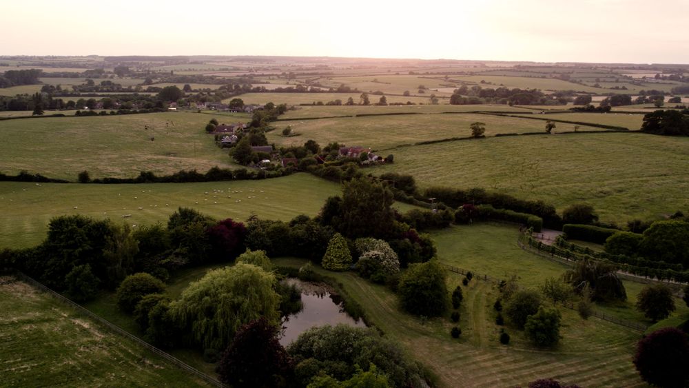 Aerial view of a sunset-lit countryside with green fields, trees, a small pond, and a few houses scattered in the distance.