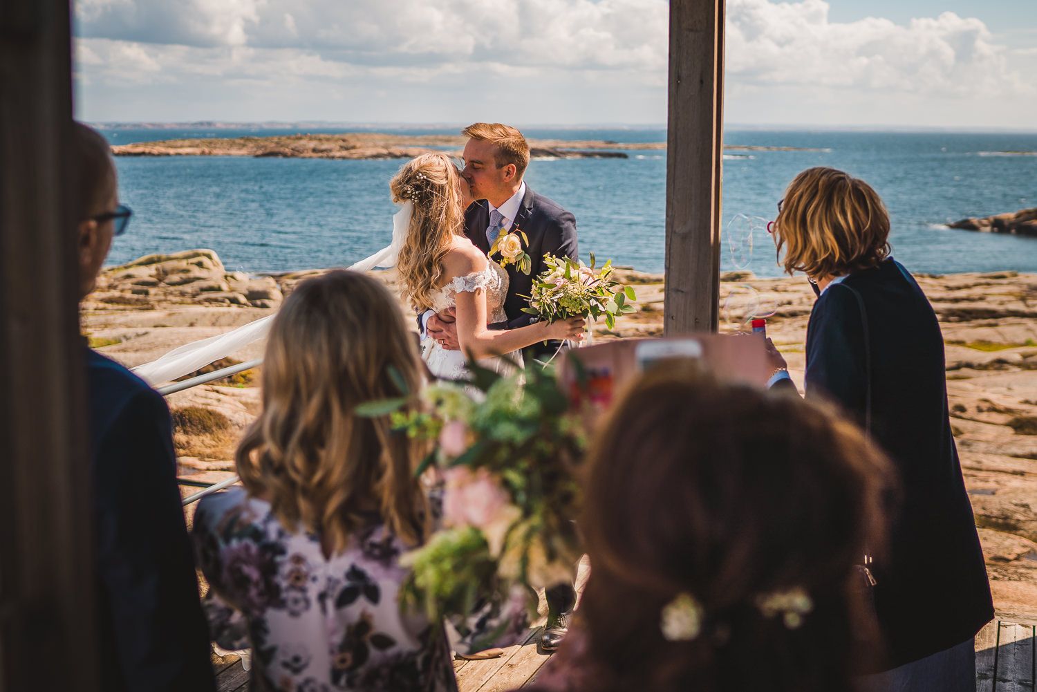 Brudpar kysser varandra utanför kapellet på Hållö en sommardag med sol och blått hav i bakgrunden