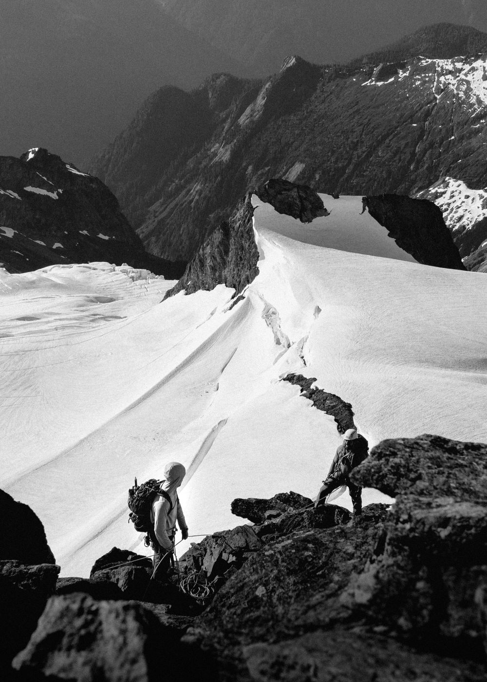 Climbers traverse a snowy mountain ridge with dramatic rocky cliffs in the background.