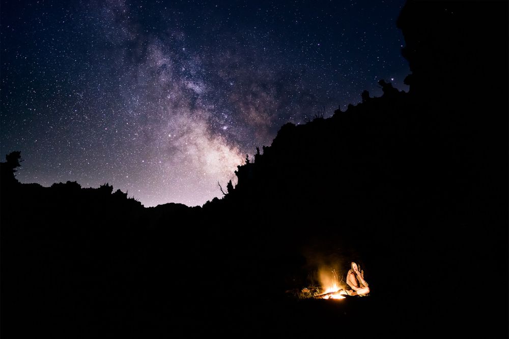Campfire glows beneath starry Milky Way sky with dark mountain silhouettes.