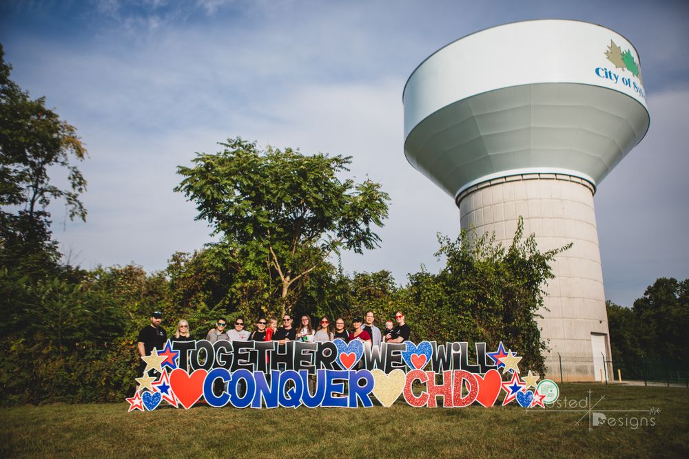 Conquering CHD organization with people behind the sign in front of a water tower.