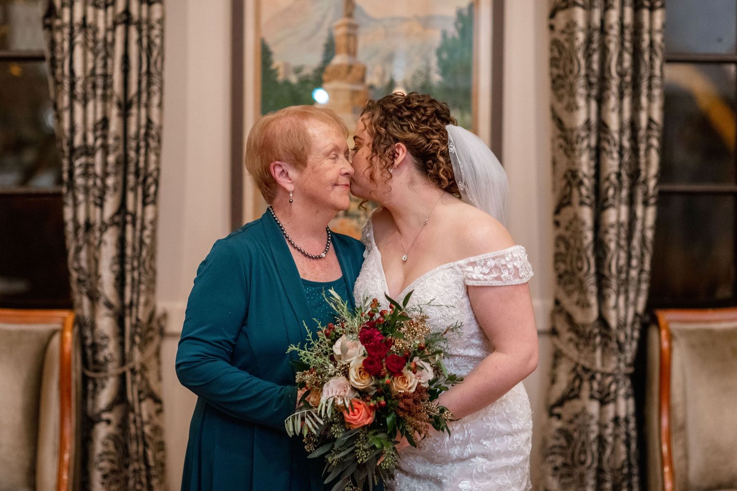 Two people embrace while holding a vibrant red and green bouquet against a curtained background.