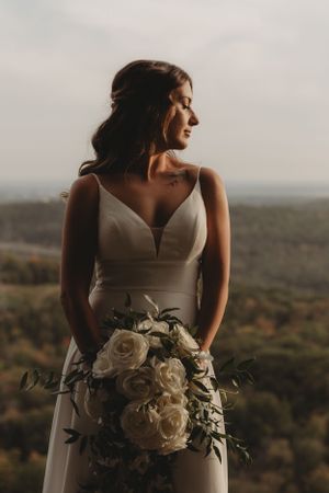Bride in elegant white dress holds bouquet of roses against mountain vista backdrop.