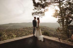 A romantic sunset wedding photo overlooking rolling hills from a wooden deck with pine trees framing the scene.