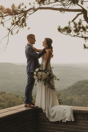 Romantic wedding couple stands on wooden deck overlooking misty mountain landscape at sunset.