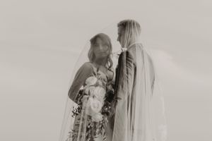 Artistic black and white portrait of couple in wedding attire against cloudy sky.