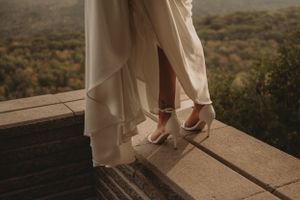 Close-up of white wedding shoes and flowing dress hem on a wooden observation deck.