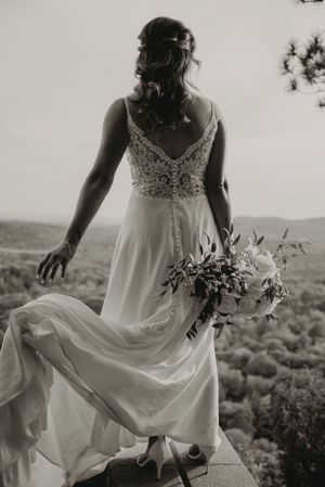Artistic black and white photo of a flowing wedding dress and bouquet against a mountain landscape.