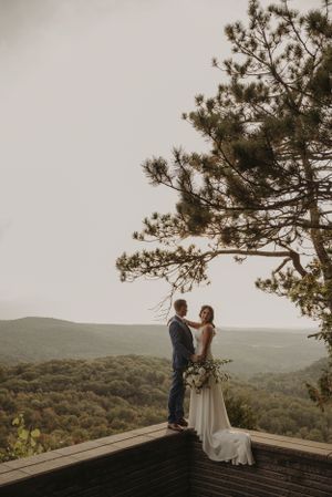 Couple stands on wooden deck overlook framed by pine branches against mountain vista.