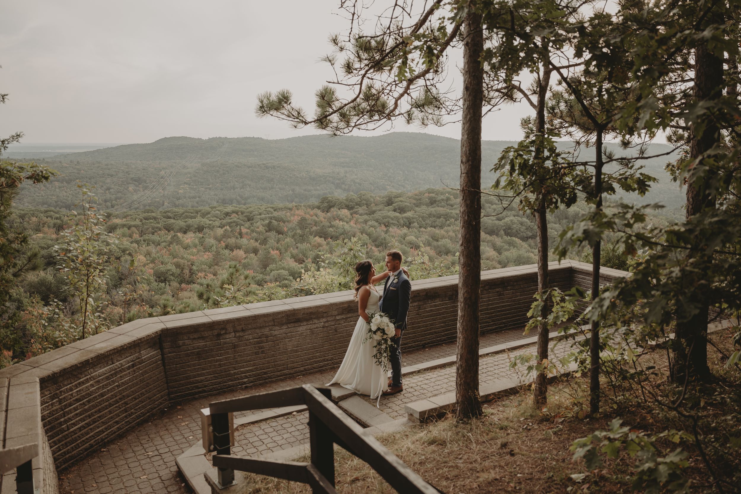 Wedding couple stands on wooden observation deck overlooking vast mountain landscape.