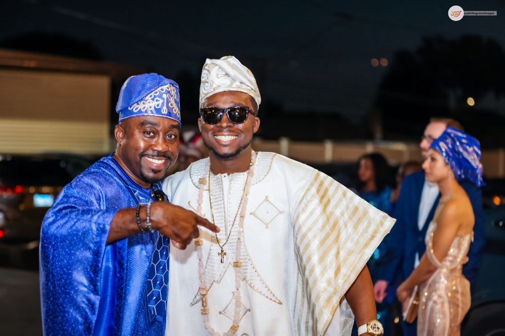 Two men in Nigerian traditional agbada outfits smiling and posing outdoors at a wedding.
