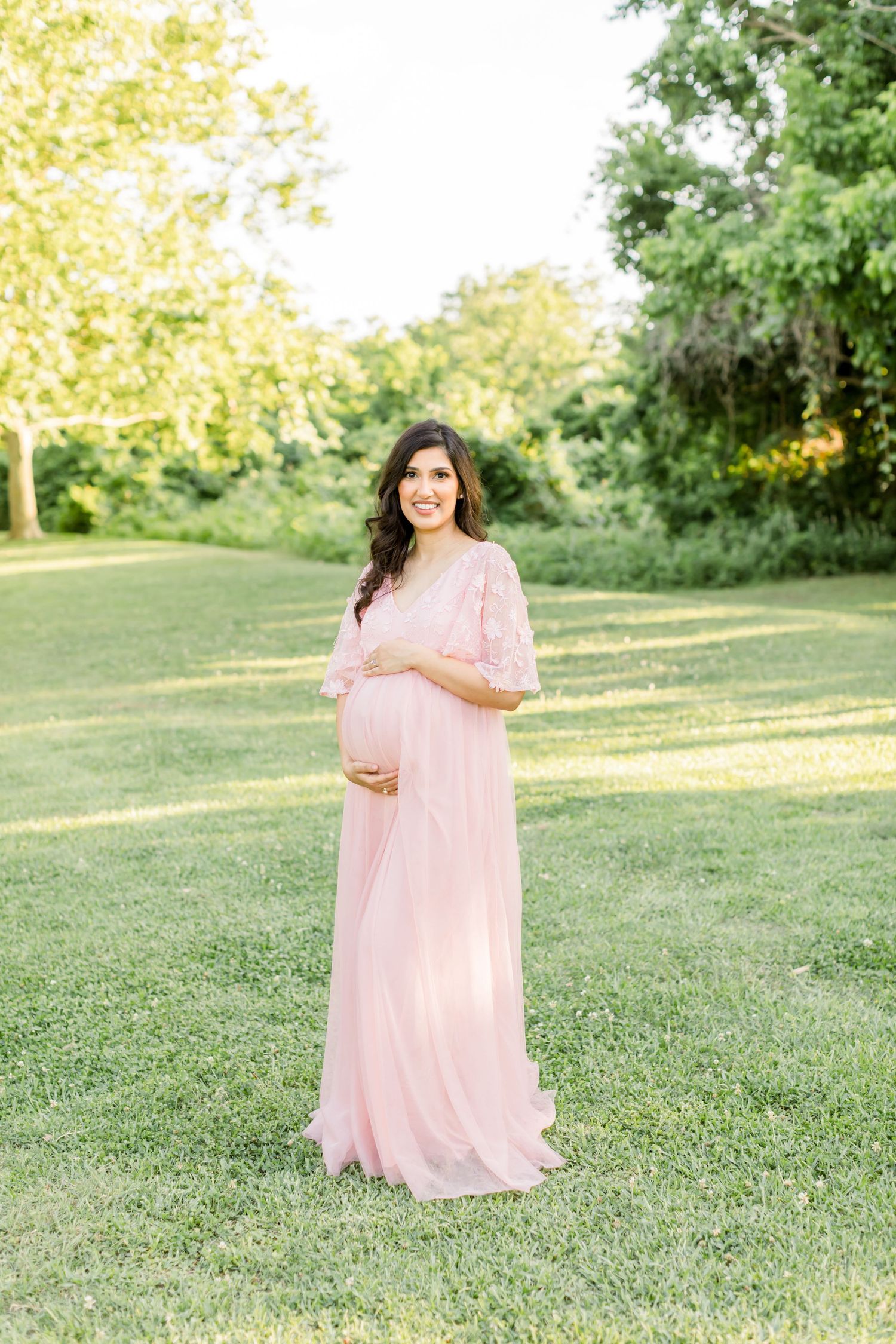 Woman in flowing pink dress cradles baby bump while standing in sunlit outdoor garden setting.