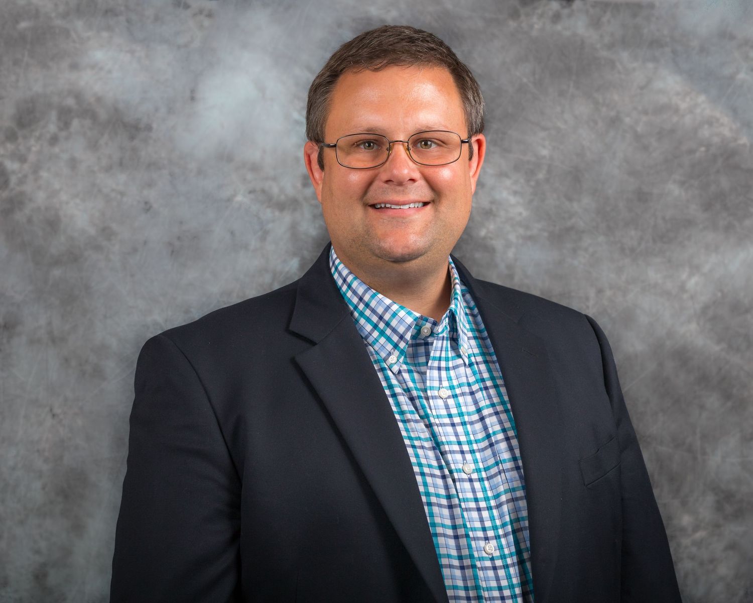 Professional business portrait in navy suit and checkered shirt against gray background.