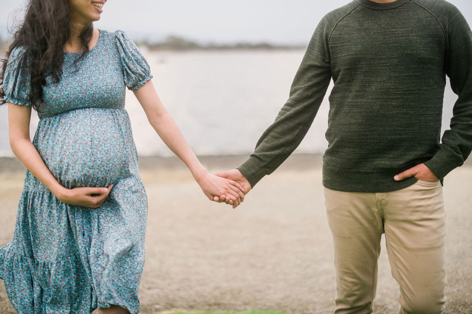 A couple holds hands by the water, with the woman wearing a flowing blue maternity dress.