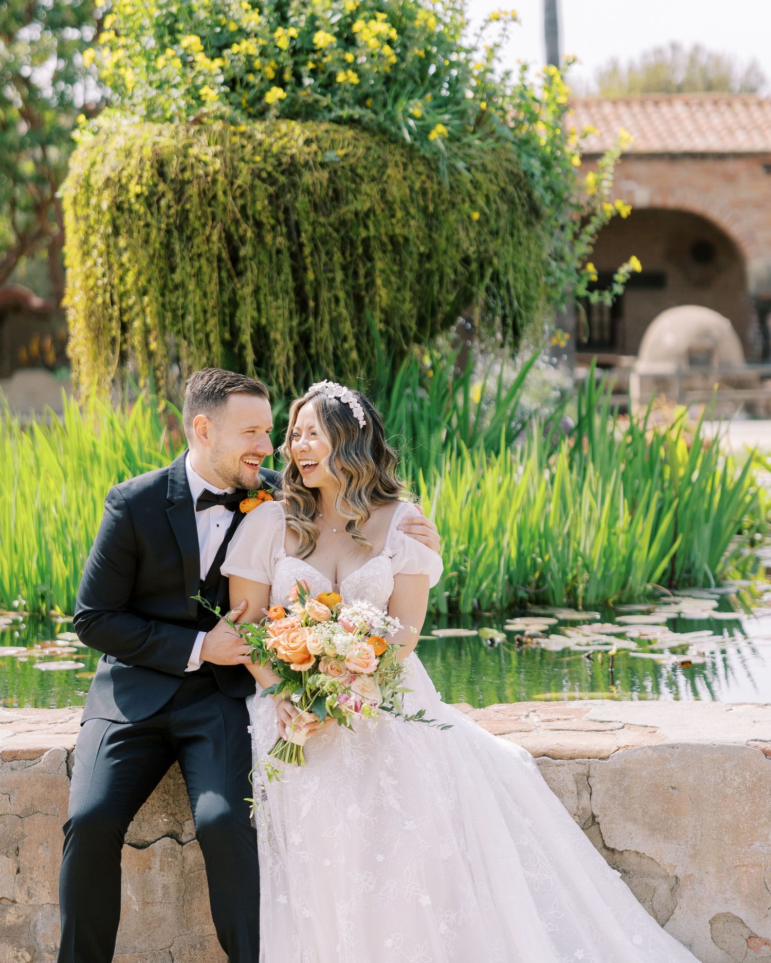 Joyful outdoor wedding portrait of a laughing bride and groom sitting on a stone wall next to a lush pond.