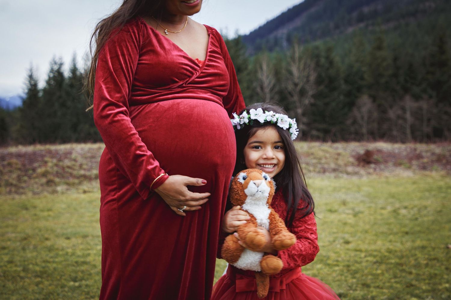 A pregnant mother holding her belly while standing near her daughter who hugs a stuffed animal