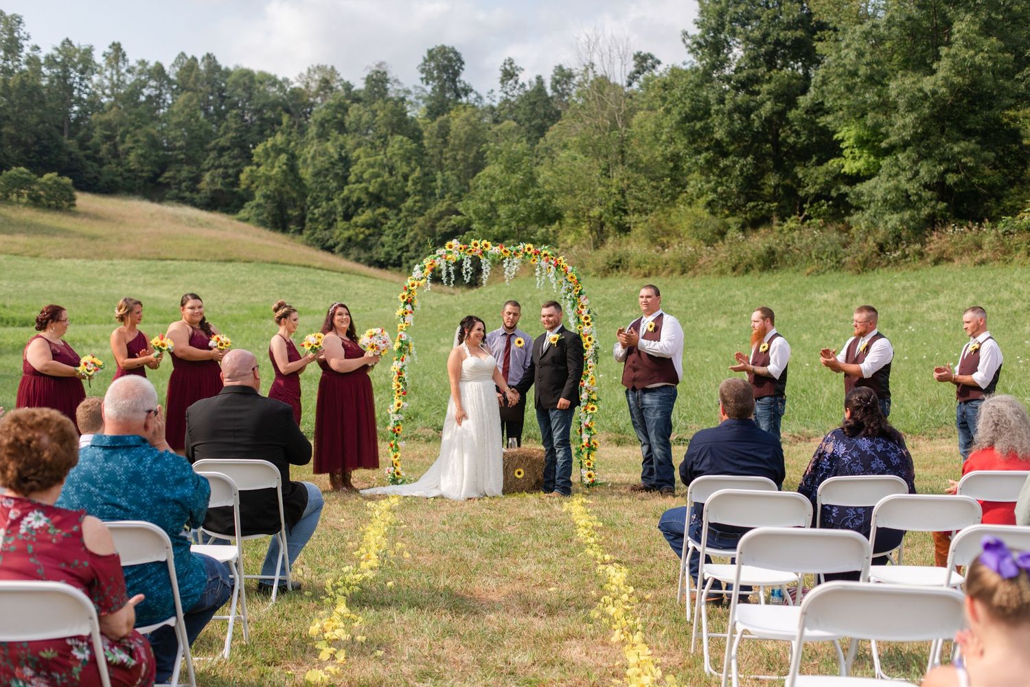 Rustic outdoor wedding setup with white chairs and sunflower arch in countryside setting.