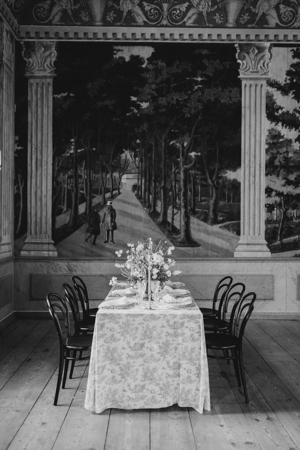 Black and white photo of wedding table setup against a classical mural featuring columns and a tree-lined path.