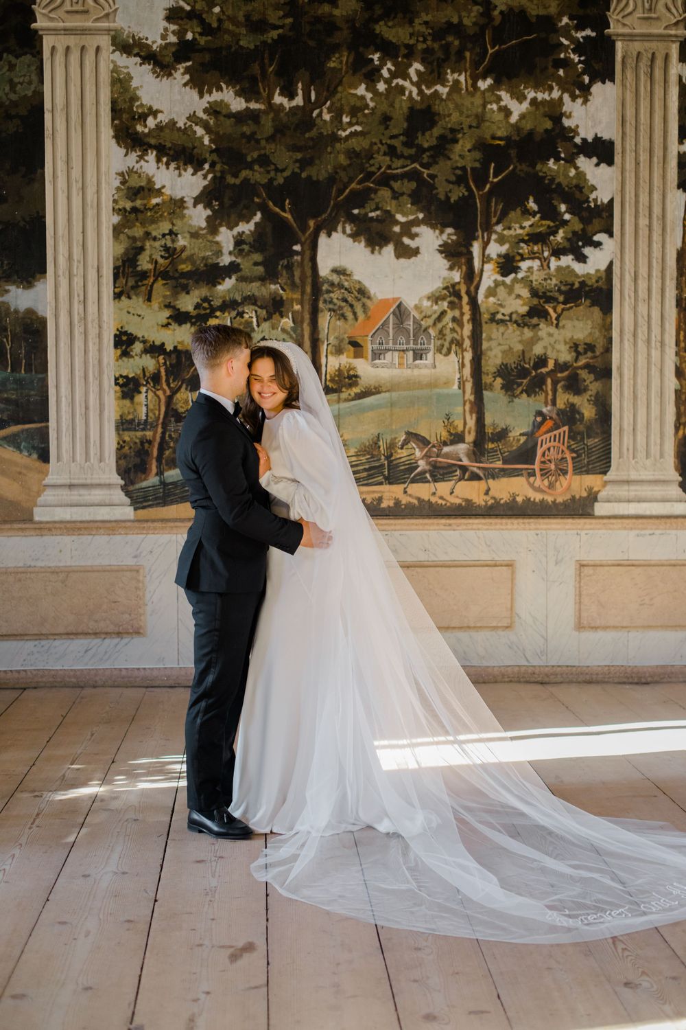 A bride and groom share an intimate moment in front of a pastoral wall mural.