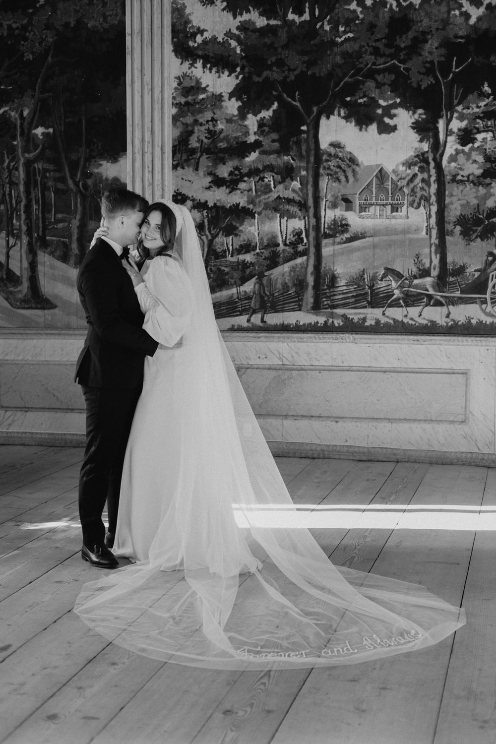 A black and white portrait of newlyweds with a long veil against a scenic mural backdrop.