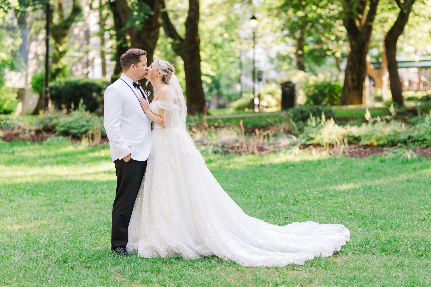 Elegant garden wedding portrait with flowing dress train in sunlit park setting.