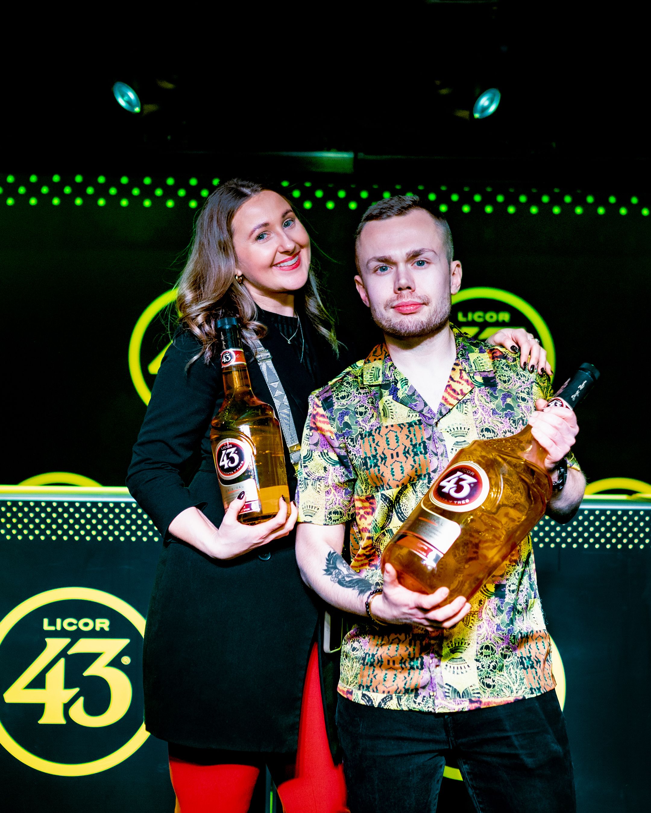 Two people holding Licor 43 bottles pose together at branded event with neon lighting.