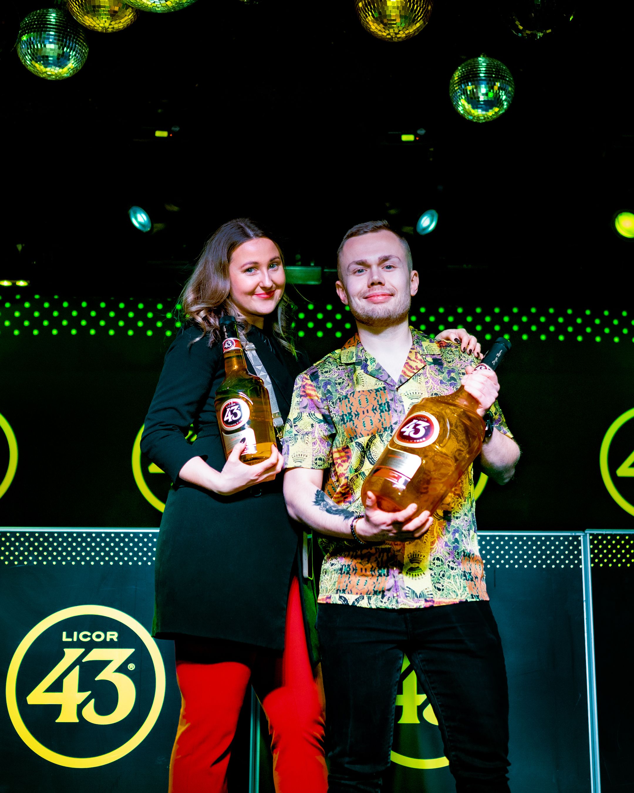 Duo displaying Licor 43 bottles at promotional event with glowing green lighting effects.