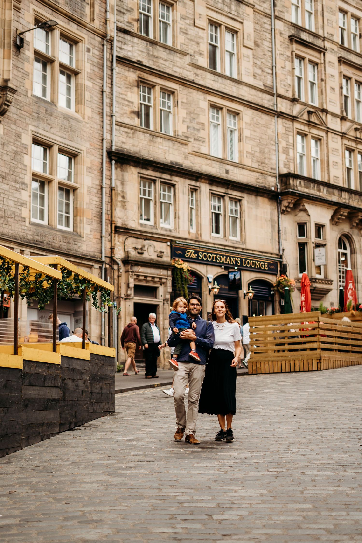 People walking past historic stone buildings along a cobblestone street in Edinburgh.