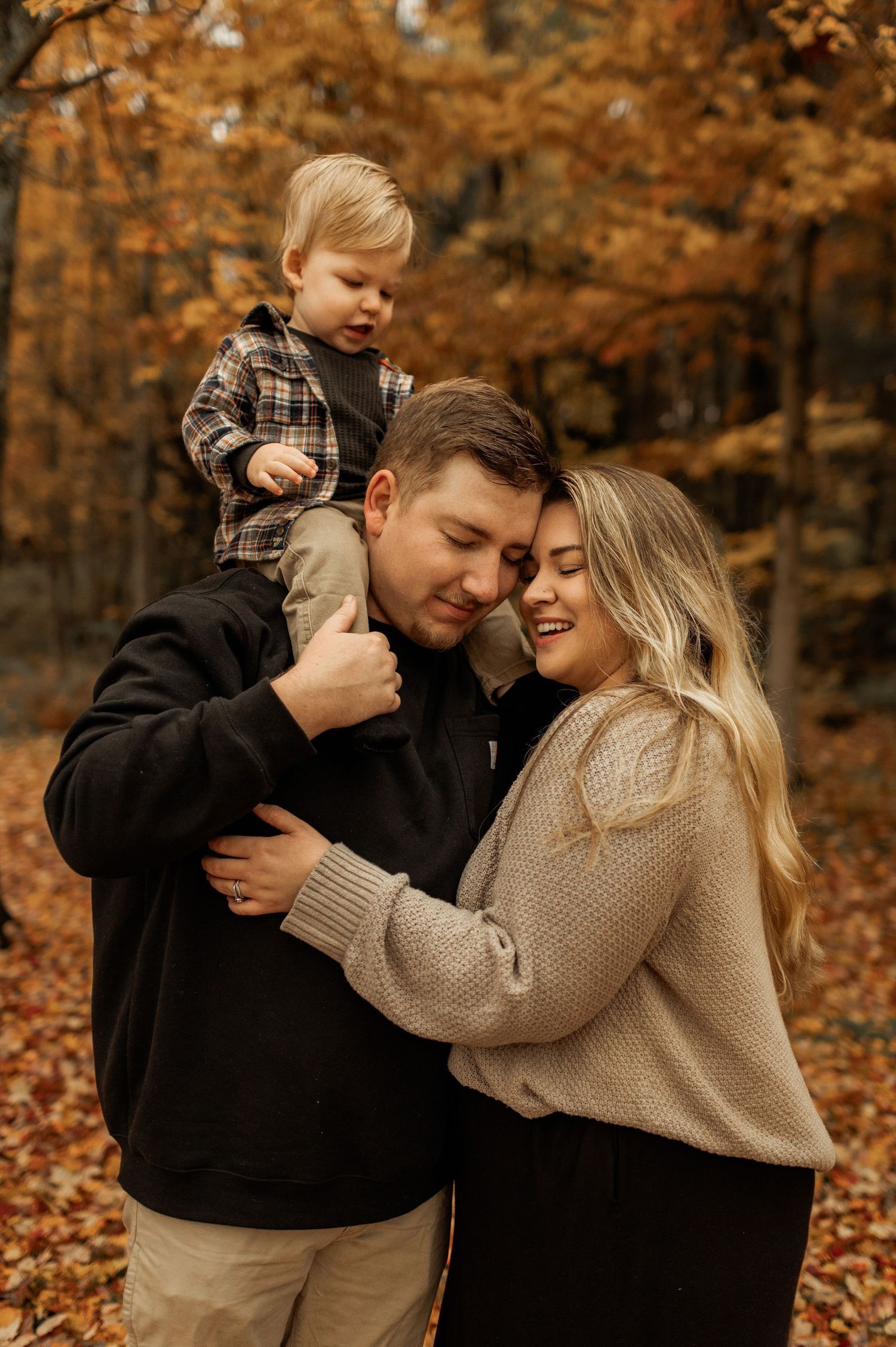 Family shares tender moment amongst fallen autumn leaves in forest setting.