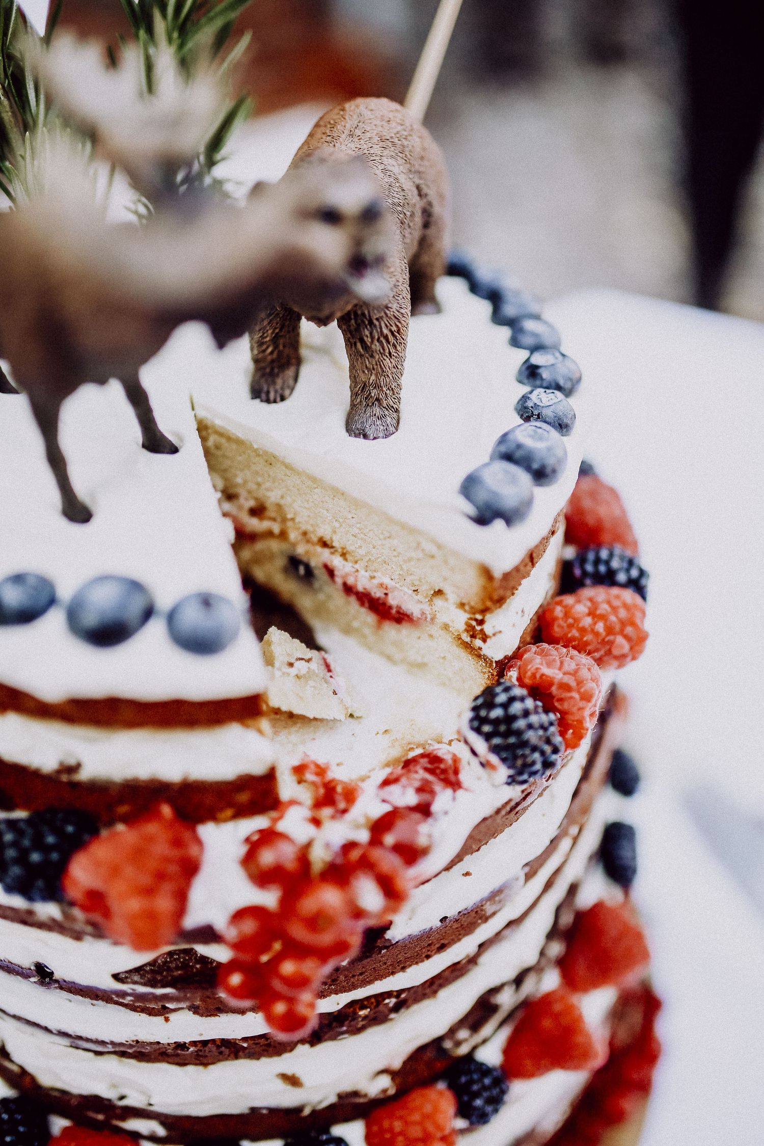 Close-up view of a rustic naked wedding cake decorated with fresh berries and small animal figurines on top.