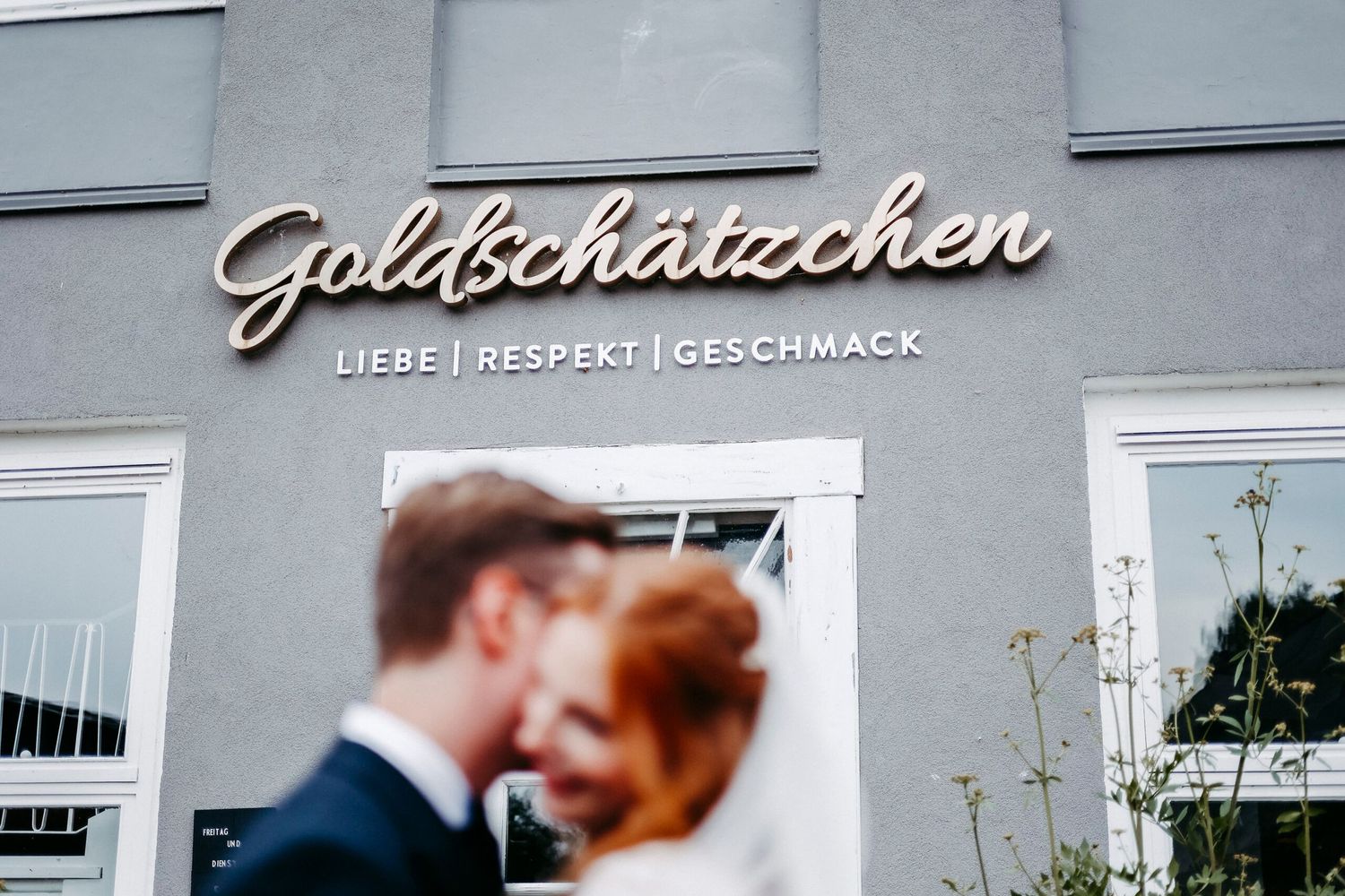 A couple kisses outside Goldschatzchen restaurant with stylish cursive signage on the building exterior.