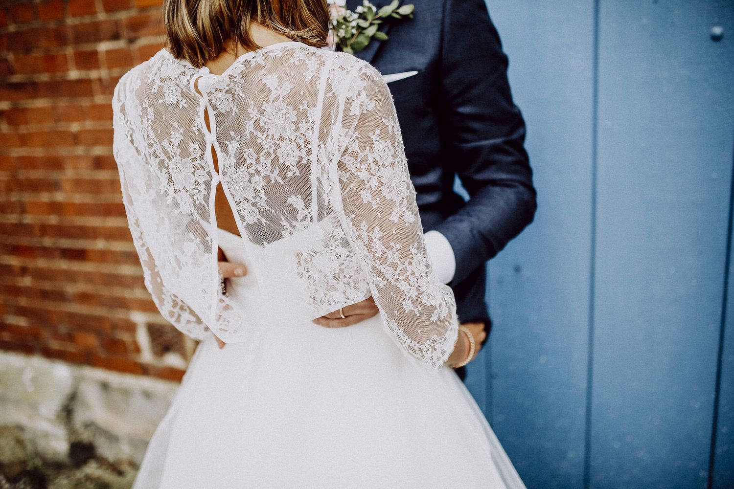 Elegant lace-back wedding dress with long sleeves against rustic brick wall and blue door.
